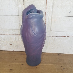 May include: A tall, sculptural vase featuring a smooth, matte finish with a gradient color transition from deep purple at the base to a lighter, muted blue at the top. The vessel has an organic, flowing shape with a narrow, asymmetrical opening.