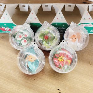 May include: Five clear spherical plastic ornaments, each containing a unique miniature scene. The scenes include white flowers, green foliage with red berries, a small figure in white, a figure in a teal hat, and a red poinsettia flower.