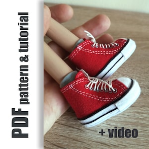 Może przedstawiać: Para miniaturowych czerwonych tenisówek z płótna z białymi sznurowadłami i podeszwami. Buty trzymane są w dłoni, z widocznym tekstem "PDF pattern & tutorial" i "+ video".