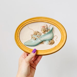Pode incluir: Uma imagem oval emoldurada com uma imagem 3D de um sapato azul com um grupo de ratos dentro. O sapato é decorado com flores e um laço. A moldura é de cor amarela clara.