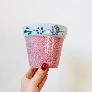 May include: A pink ceramic flower pot with a white rim and a painted floral design in blue, yellow, and pink.
