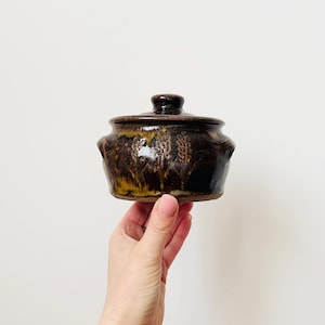 May include: A small, brown ceramic pot with a lid. The pot has a floral design and two handles.