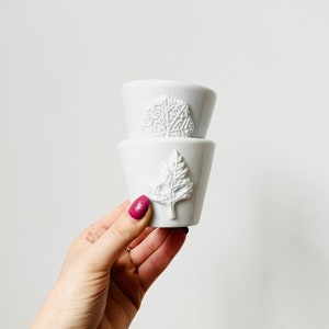 May include: Two white ceramic cups with a tree design. The cups are stacked on top of each other.