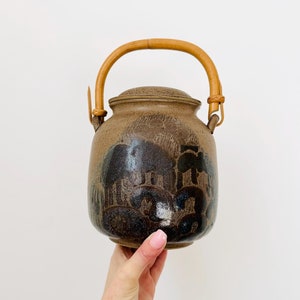 May include: A brown ceramic teapot with a bamboo handle and a black and white design. The teapot has a lid and is decorated with a pattern of stylised trees and clouds.