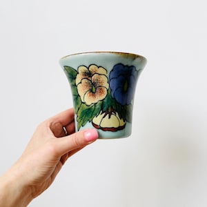 May include: A light blue ceramic vase with a floral design. The vase features yellow, blue, and cream-coloured flowers with green leaves. The vase is held by a hand against a white background.