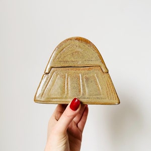 May include: A ceramic box with a lid, shaped like a mountain. The box is a light brown colour with a speckled glaze.
