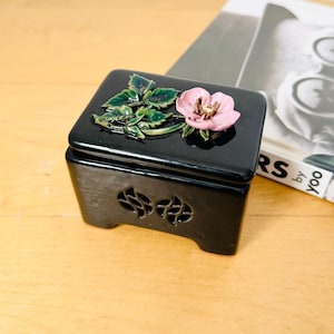 May include: A black ceramic box with a floral design. The box has a lid with a raised pink flower and green leaves. The box has a decorative design on the front and four small feet.