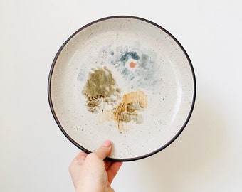 May include: A white ceramic plate with a brown rim and a speckled glaze. The plate features a hand-painted design of a landscape with trees, clouds, and a sun.
