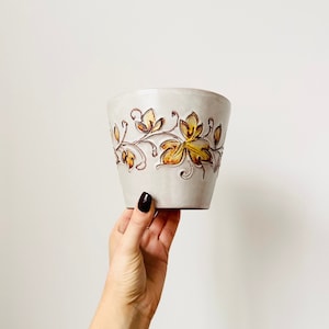 May include: A white ceramic planter with a brown and yellow floral design. The planter is decorated with a repeating pattern of stylised leaves and flowers.