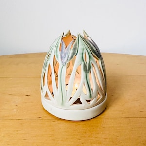 May include: White ceramic candle holder with a delicate floral design. The holder is shaped like a dome with cutouts that resemble leaves and flowers. The candle holder is lit and the light shines through the cutouts.