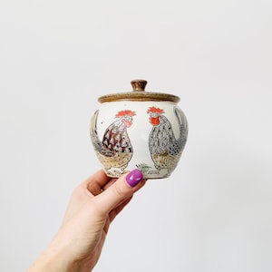 Puede incluir: Un tarro de cerámica blanco con tapa marrón y diseño de gallo. El tarro tiene dos gallos pintados en él, uno a cada lado. Los gallos son negros, marrones y rojos.
