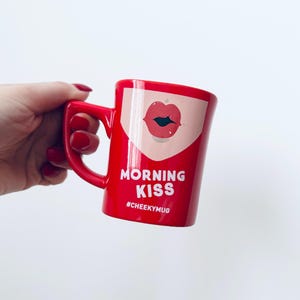 Puede incluir: Una taza de cerámica roja con asa, con un gráfico de labios rojos y las palabras "MORNING KISS" en blanco. La taza también tiene el hashtag "#CHEEKYMUG" impreso. La taza es de color rojo vibrante.