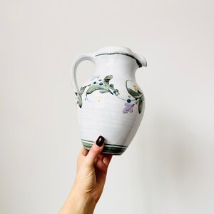 May include: White ceramic pitcher with a green, purple, and yellow floral design. The pitcher has a handle and a spout.