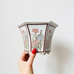 May include: A white ceramic planter with a hexagonal shape and a floral design. The planter has a brown rim and features pink and blue flowers with green leaves.
