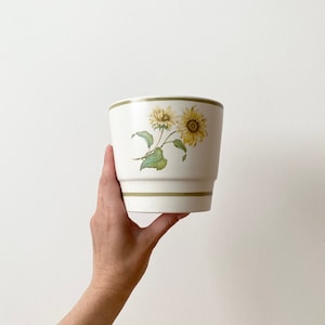 Op de afbeelding: Witte keramische pot met een groene rand en een zonnebloemdessin. De pot wordt in een hand gehouden.