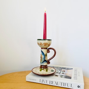 Puede incluir: Un portavelas de cerámica con un diseño floral azul y verde y un asa. El portavelas está sobre un libro titulado "Live Beautiful" de Caldeone. Una vela rosa está encendida en el portavelas.