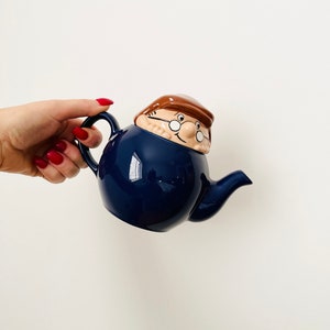 May include: A blue ceramic teapot with a cartoon face on the lid. The teapot has a brown hat and glasses.