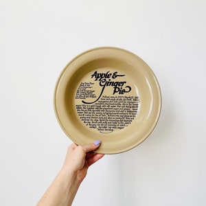 Puede incluir: Fuente de tarta de cerámica color canela con las palabras "Apple & Ginger Pie" en escritura negra. La fuente presenta una receta impresa en texto negro alrededor del borde. La fuente está sostenida por una mano.