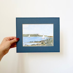 Puede incluir: Una pintura de acuarela enmarcada de un paisaje mar&iacute;timo con tres veleros en la distancia. La pintura est&aacute; ambientada en un cielo azul y una costa verde y herbosa.