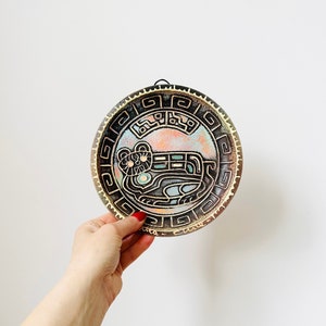 May include: A round ceramic wall plate with a black and gold design. The plate features a stylised animal, possibly a jaguar, with intricate details. The plate has a hanging loop on the top.