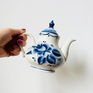 Può includere: Una teiera bianca con un motivo floreale blu. La teiera ha un coperchio con un pomo in cima. La teiera è dipinta a mano con fiori e foglie blu.