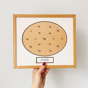May include: A framed display of vintage trout flies, arranged in a circle on a tan background. The frame is made of light wood and has a white mat. The text "Trout Flies circa - 19th century" is printed on a white label below the circle.