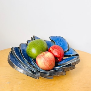 May include: A blue ceramic bowl with a unique, abstract design. The bowl is filled with three pieces of fruit: a green apple and two red apples.