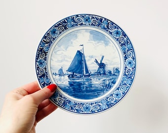 May include: A round, blue and white decorative plate featuring a nautical scene with a sailboat and windmills. The plate has a floral border design. The plate is held by a person with red nail polish.
