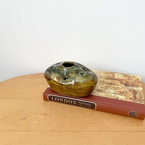 May include: A green and brown ceramic bud vase with a rounded shape and multiple holes for flowers. The vase is sitting on a book titled "London Portrait of a City".