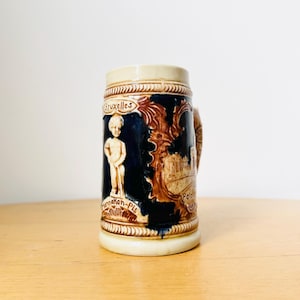 Op de afbeelding: Een klein, keramisch bierglas met een blauwe achtergrond en bruine en witte details. Het glas toont een afbeelding van een gebouw en een figuur, samen met de tekst "Bruxelles" en "Manneken-Pis".