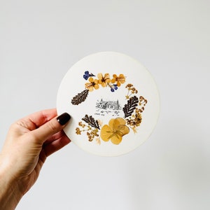 May include: A white circular card with a pressed flower design. The flowers are yellow, brown, and blue. The card has a black and white illustration of a house with the text "RANDS END" underneath.