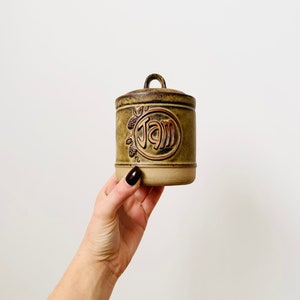 May include: A brown ceramic jar with a lid and a brown and orange "JAM" inscription. The jar has a floral design around the inscription.