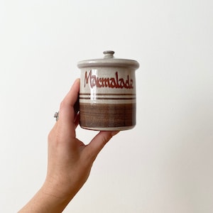 Può includere: Un barattolo di ceramica bianco con strisce marroni e la parola "Marmalade" scritta in rosso. Il barattolo ha un coperchio ed è tenuto da una mano.