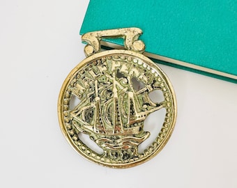 Op de afbeelding: Een gouden metalen boekenlegger in de vorm van een schip met een gedetailleerd ontwerp. De boekenlegger heeft een ronde vorm met een verhoogde rand en een decoratieve rand.