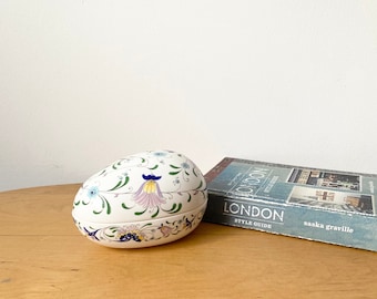 May include: A white ceramic egg-shaped trinket box with a floral design in shades of blue, green, pink, and yellow. The box is closed and sitting on a wooden surface.