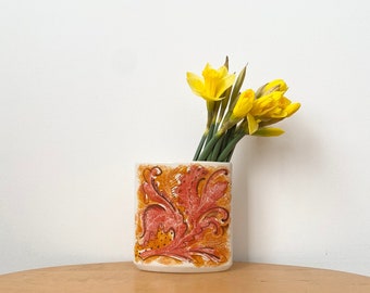 Könnte beinhalten: Eine weiße Keramikvase mit einem roten und orangefarbenen Blumendesign, die einen Strauß gelber Narzissen hält.