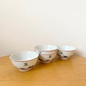 Può includere: Quattro ciotole in ceramica bianca con un bordo rosso e disegni floreali verdi e rossi. Le ciotole sono su una superficie di legno marrone chiaro.