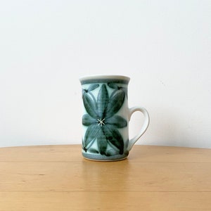 Könnte beinhalten: Eine weiße Keramiktasse mit grünem Blumendesign. Die Tasse hat einen Henkel und steht auf einer Holzoberfläche.
