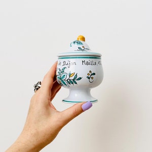 Puede incluir: Tarro de cerámica blanca con un diseño floral verde y amarillo. El tarro tiene una tapa y una base. El tarro está etiquetado con "Maille Dijon" en letras negras.