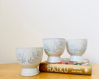 Könnte beinhalten: Drei weiße Keramikbecher mit blauen Blumendesigns, gestapelt auf einem Buch mit dem Titel "HAIKU", herausgegeben von Jackie Hardy.