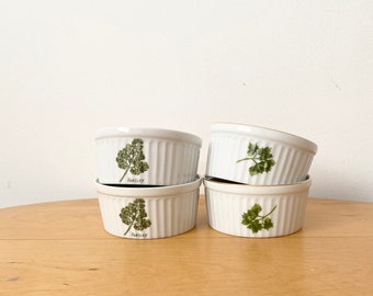 May include: Four white ceramic ramekins with a green parsley design and the word "Parsley" printed on the bottom. The ramekins are stacked on a light brown wooden surface.