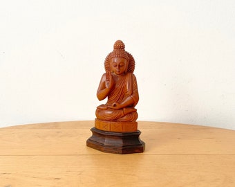 May include: A wooden statue of Buddha sitting in a meditative pose with his right hand raised in the Abhaya mudra, a gesture of reassurance and protection. The statue is carved in a reddish-brown wood and has a detailed face and intricate clothing.