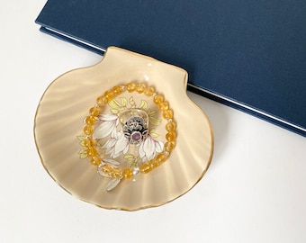 Puede incluir: Un pequeño plato de cerámica blanco en forma de concha de mar con un diseño floral. Un brazalete de piedras amarillas con un dije de plata descansa dentro del plato.