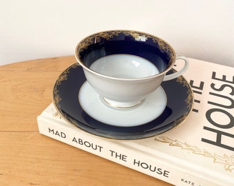 May include: A white teacup and saucer with a blue and gold rim. The saucer has a gold floral design. The teacup and saucer are sitting on a book titled "Mad About The House" by Kate Watson-Smyth.
