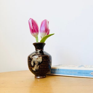 Op de afbeelding: Een kleine, donkerbruine en witte keramische vaas met een getextureerd oppervlak bevat twee roze tulpen. De vaas staat op een licht houten tafel naast een boek getiteld "The Little Book of Gods" van Meik Wiking.