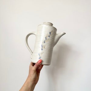 May include: A white ceramic coffee pot with a blue floral design. The pot has a tall, cylindrical shape and a spout on the side.