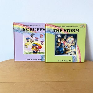 May include: Two children's books with rainbow covers. The first book is titled "Scruffy" and the second book is titled "The Storm". Both books are part of the "Adventures of the Rainbow Scarecrows" series.