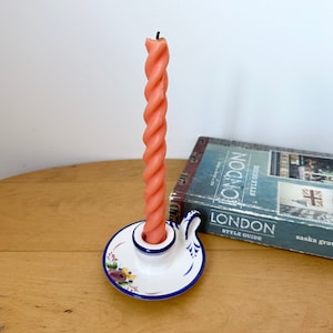 Op de afbeelding: Een witte keramische kaarsenhouder met een blauw en bloemig ontwerp. De houder heeft een gedraaide oranje kaars erin. De kaarsenhouder staat op een houten tafel naast een boek met de titel "London Style Guide".