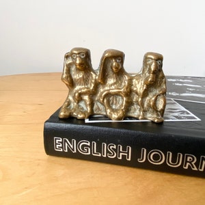 May include: Three brass monkey figurines sitting side by side. The monkeys are in a seated position with their hands clasped together. The monkeys are all facing forward. The figurines are on top of a black book with the title "ENGLISH JOURNAL" printed in white.
