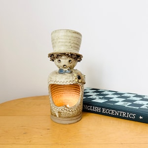 Puede incluir: Un portavelas de cerámica con forma de persona que lleva un sombrero. El portavelas tiene una superficie texturizada y un pequeño agujero en la parte delantera para una vela. La vela está encendida y brillando.